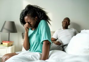 A heartbroken Black woman sits on the edge of a bed crying, while a man sits behind her looking away, depicting emotional pain, relationship conflict, and the need to guard your heart after heartbreak.